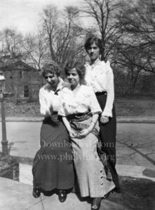 Black-and-white photo of three women posing in front of a road and a house