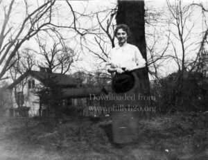 Black-and-white photo of a young woman standing in front of a large tree and a small house