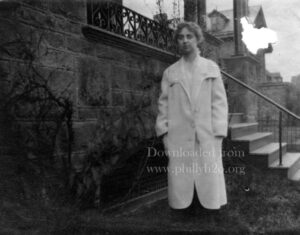 Black-and-white photo of young woman in white coat in front of large house