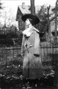 Black-and-white photo of a young woman wearing a large coat and black hat in front of a house with a yard.