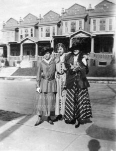Black-and-white photo of three women standing on a sidewalk in front of a row of houses.