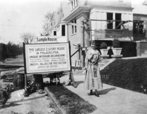 Black-and-white photo of a young woman in a long dress standing next to a sign in front of a house.