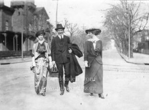 Black-and-white photo of a man with a mustache and two girls in dresses and hats walking through an empty intersection.