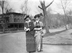 Black-and-white photo of two young women standing in the street in front of a large house and many trees