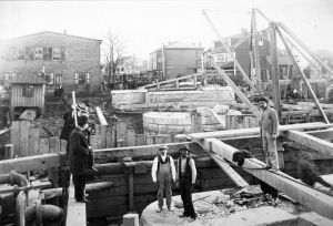 Black and white photo of two men facing the camera and several facing away while standing and sitting on a board