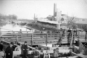 Black and white photo of several men in the foreground, some wearing hats, and a few others standing behind them on a wooden bridge