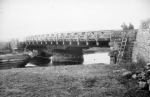 Black and white photo showing a wooden truss bridge over a creek