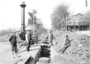 men standing around a trench with pipes seen crossing through it. there is a steam engine on wheels behind them to the left, and a building in the background on the right with a sign they says Hotel Abbey, T G Schmidt & Sons, Pilsner & Puritan Beer.