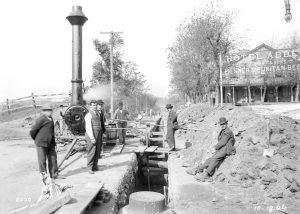 men standing around a trench with pipes seen crossing through it. there is a steam engine on wheels behind them to the left, and a building in the background on the right with a sign they says Hotel Abbey, T G Schmidt & Sons, Pilsner & Puritan Beer.