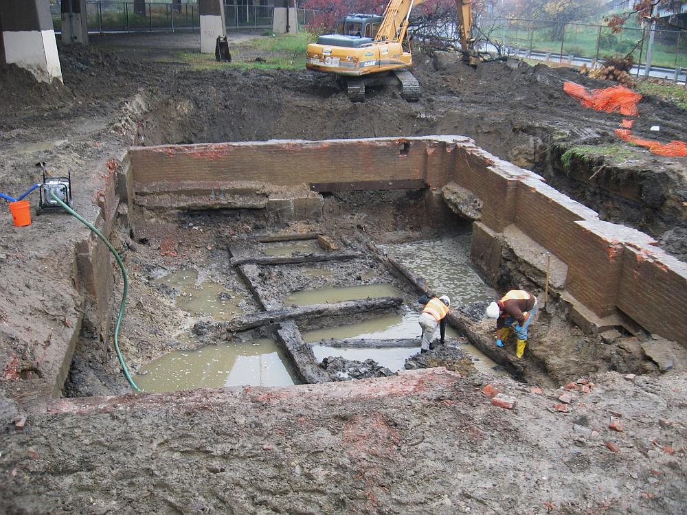 Image shows several wooden poles in a square muddy pit with wooden walls