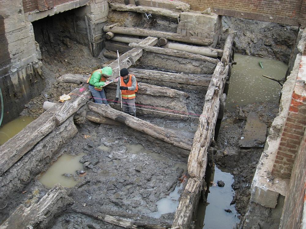 Image shows several old wooden pipes in mud and two men in protective clothing