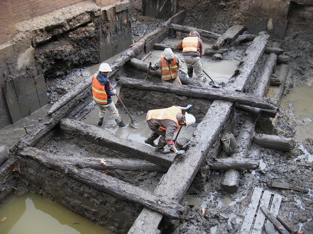 Image shows several workers in a muddy pit and several large wooden poles