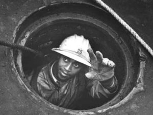 Black and white photo of a worker with darker skin, wearing protective clothing, gloves, and a hard hat with the number 25 on the front, reaching up out of a sewer manhole, only head, shoulders, and left arm visible.