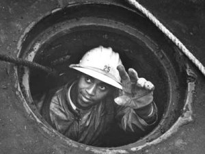 Black and white photo of a worker with darker skin, wearing protective clothing, gloves, and a hard hat with the number 25 on the front, reaching up out of a sewer manhole, only head, shoulders, and left arm visible.