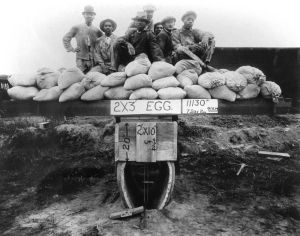 Six people and dozens of sandbags performing a load test on a concrete pipe, 1914