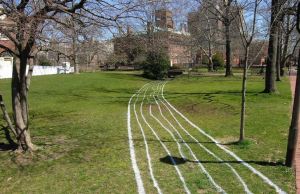 Color image of a grassy field with trees and buildings surrounding it and white lines drawn on the grass mimicking the path of a creek.
