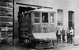 Black-and-white image shows a trolley car emerging from a building with men standing alongside