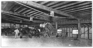 Black-and-white photo of men, horses and a cart inside of a barn-like structure