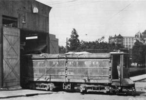 Black-and-white photo of an ash car, side view, in the doorway of a barn with trees behind