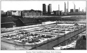 Black-and-white photo of industrial tank installation with smokestacks and factories in the background
