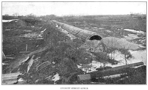 Black-and-white photograph depicts a large brick sewer tube lying on a barren landscape