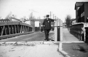 Black and white photo of a man wearing a hat and standing on a bridge with his arm on a post