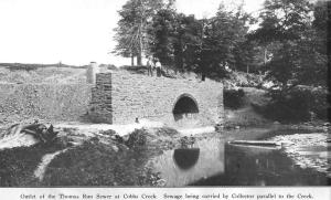 Black-and-white image of a brick sewer outlet that drains into a creek with trees and a hill behind it